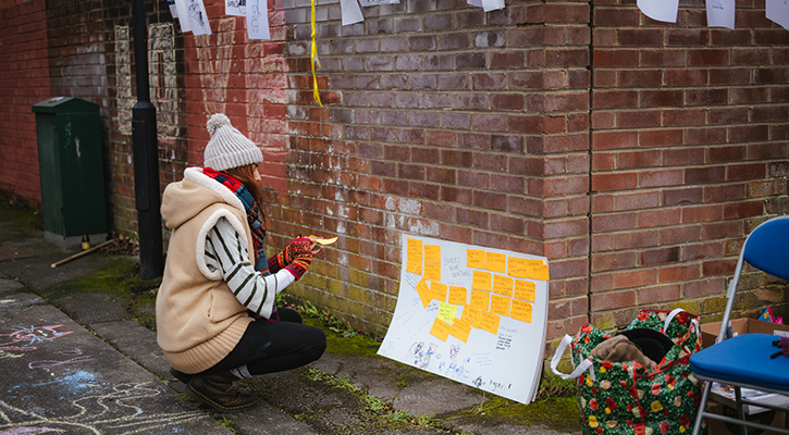 A woman in a coat and woolly hat sits in an alleyway and writes a contribution to a community feedback wall.